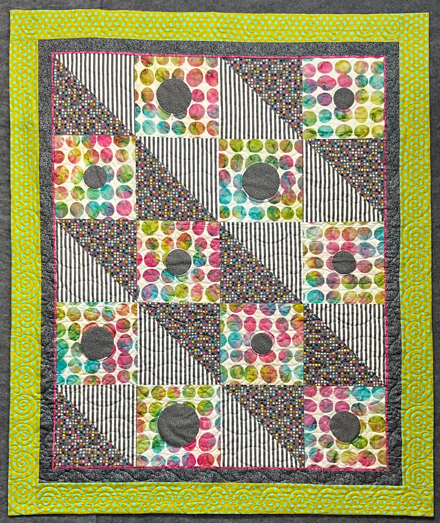 The Seeing Spots: Quilted Wall Art by Mary Cantore features colorful, diagonally divided squares with polka dot, striped, and spotted patterns, gray circular patches, a yellow-green border, and a purple stripe—perfect for a child’s room.