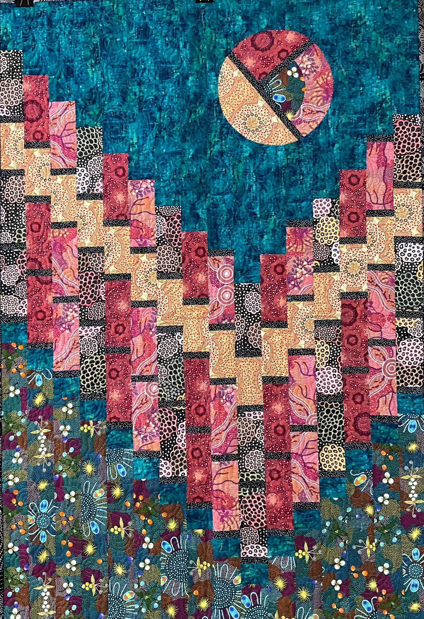 Moon Over Magdalaina: Quilted Wall Art by Mary Cantore features a vibrant sun in a teal sky above patterned pink, yellow, and red blocks resembling the Australian outback, with a floral design accenting the bottom.
