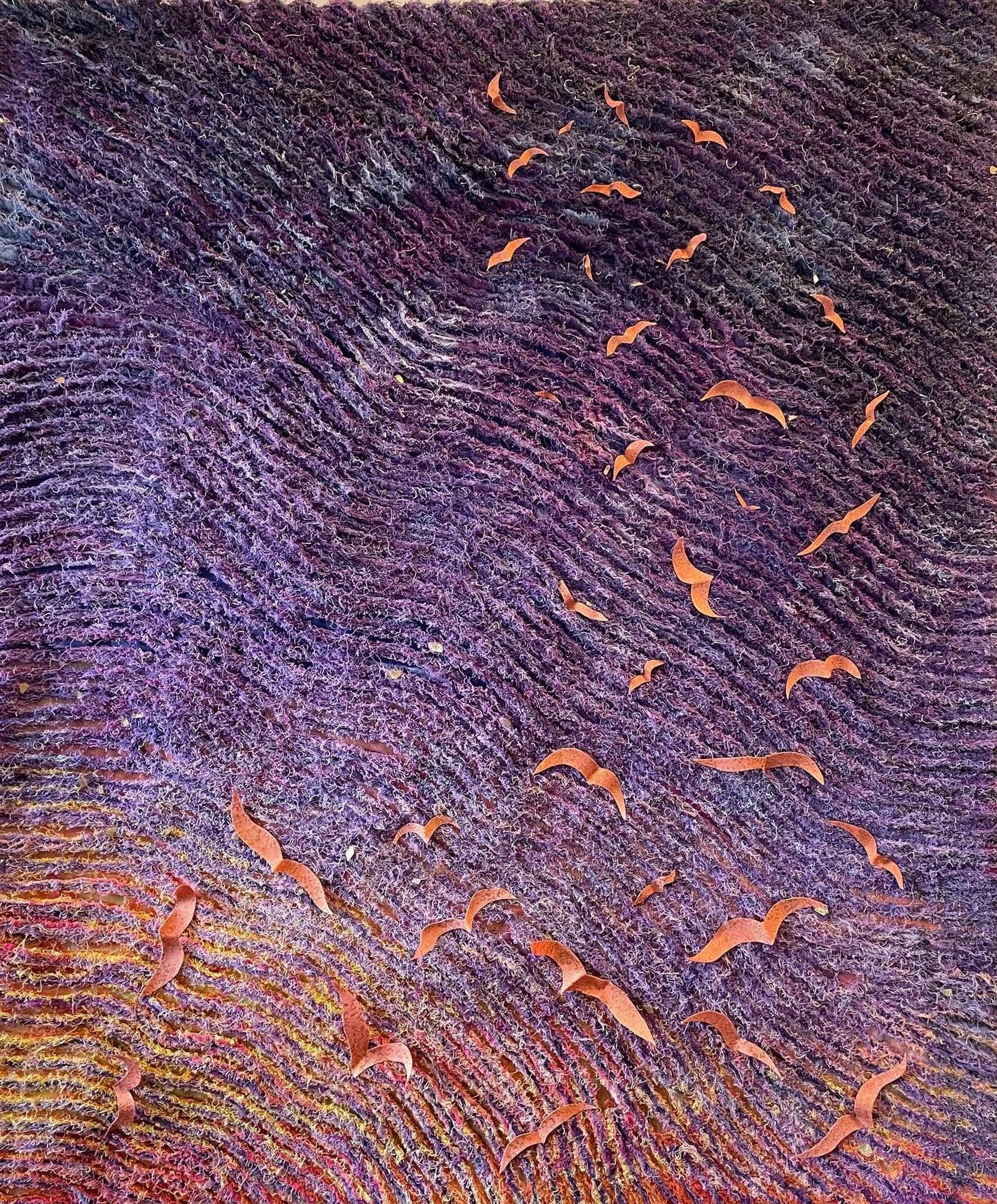 The Carry the Light wall hanging by Miki Howell features undulating purple and red waves, scattered with orange brocade birds resembling origami. Crafted from hand-dyed fabrics, it evokes movement and depth against a dark background.