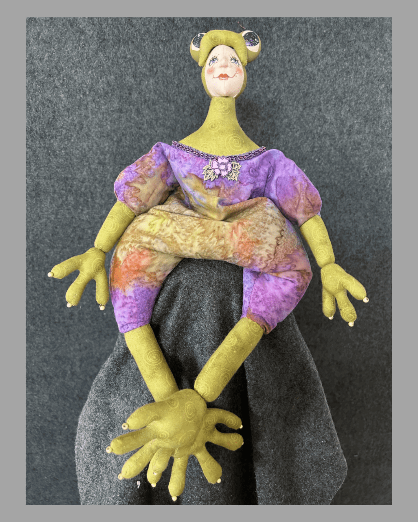 Frogissimo: Art Doll by Mary Cantore features a hand-painted face, oversized yellow hands and feet, and a purple-yellow outfit, all crafted in fabric. This whimsical collectible sits playfully against a gray fabric backdrop.