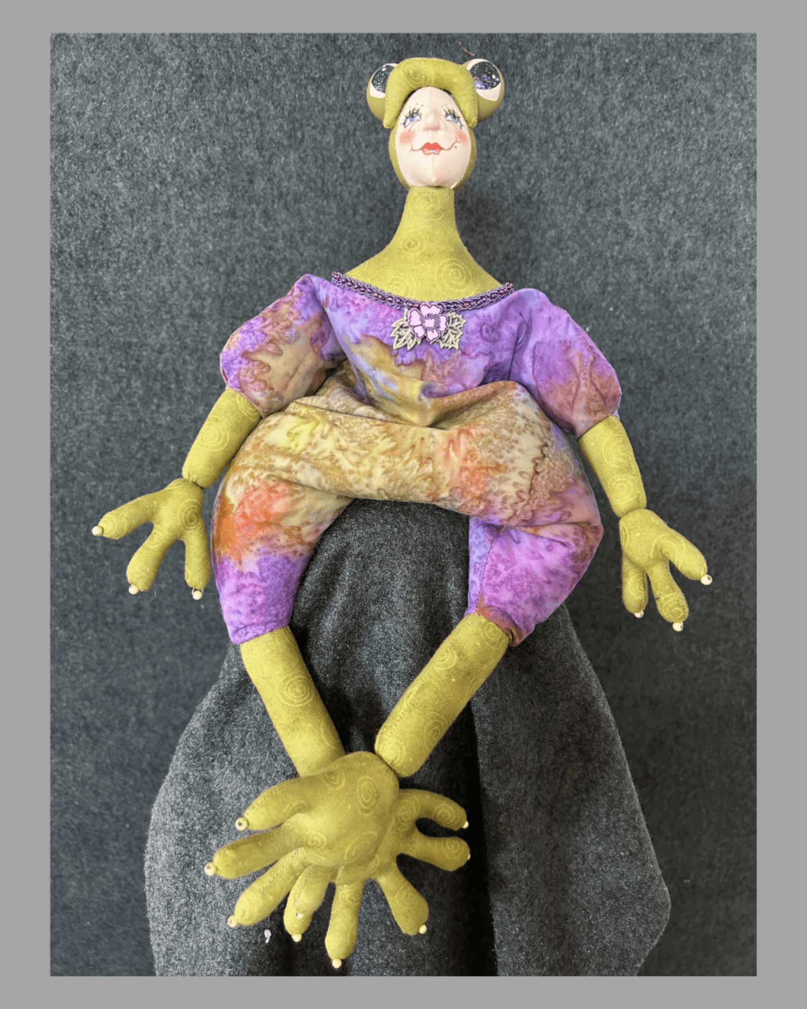 Frogissimo: Art Doll by Mary Cantore features a hand-painted face, oversized yellow hands and feet, and a purple-yellow outfit, all crafted in fabric. This whimsical collectible sits playfully against a gray fabric backdrop.