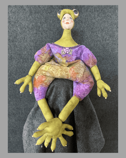 Frogissimo: Art Doll by Mary Cantore features a hand-painted face, oversized yellow hands and feet, and a purple-yellow outfit, all crafted in fabric. This whimsical collectible sits playfully against a gray fabric backdrop.
